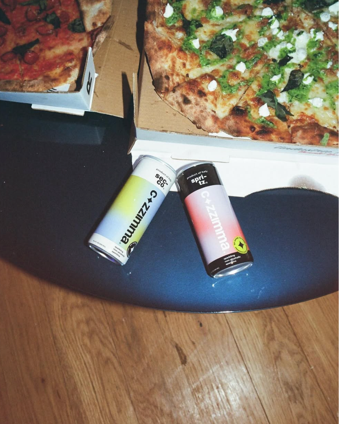 Two alcohol-free drink cans on a table with pizzas in the background